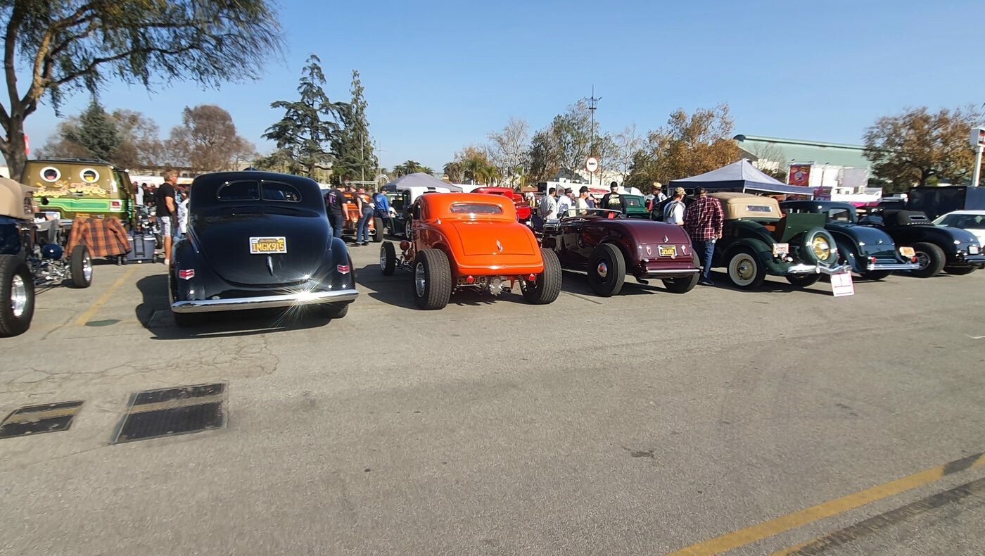 Rząd hot rodów na zlocie – czarne coupe, pomarańczowy roadster i tłum przy stoiskach