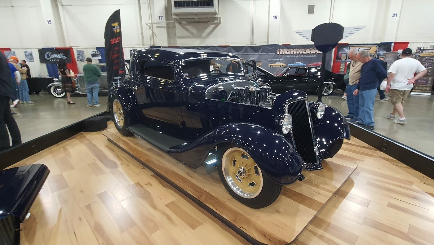 Granatowy Ford 3-window coupe z 1934 roku na złotych felgach – hot rod w błotnikach na drewnianej podłodze hali