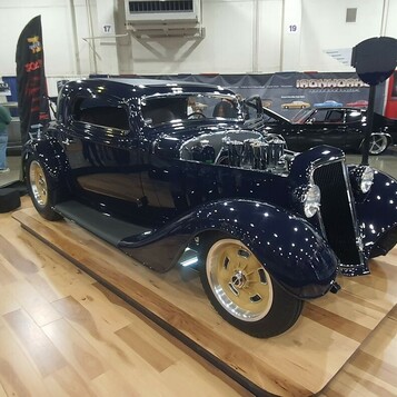 Granatowy Ford 3-window coupe z 1934 roku na złotych felgach – hot rod w błotnikach na drewnianej podłodze hali