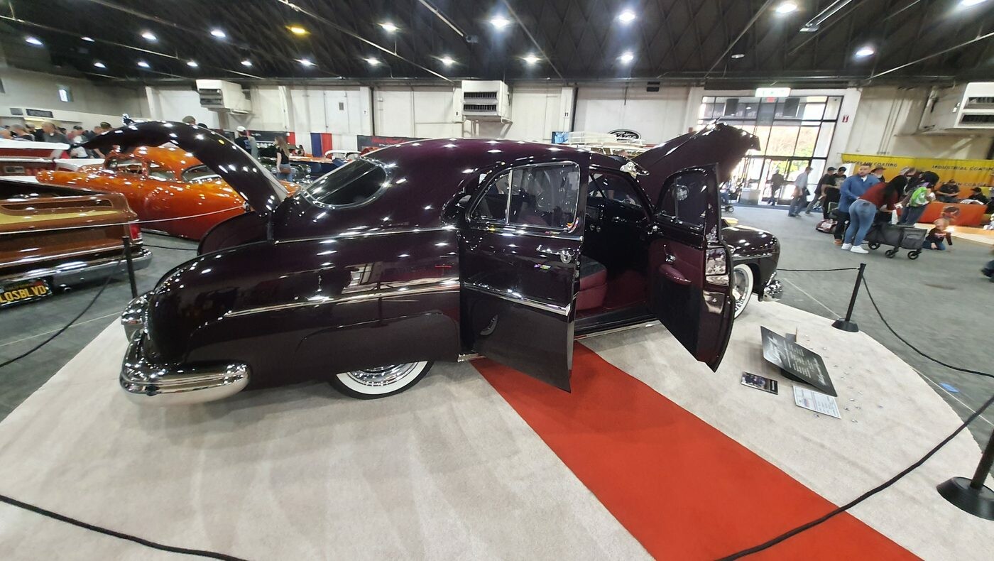 Ciemnośliwkowy kustom z lat 40. z otwartymi drzwiami i maską – lead sled na czerwono-białym podłożu w hali