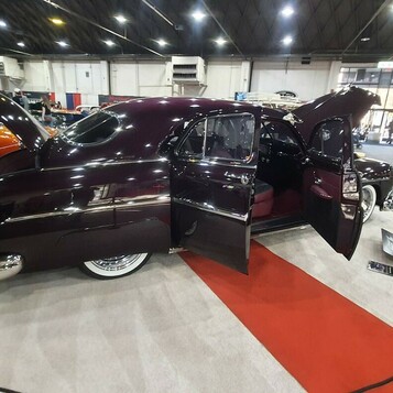 Ciemnośliwkowy kustom z lat 40. z otwartymi drzwiami i maską – lead sled na czerwono-białym podłożu w hali