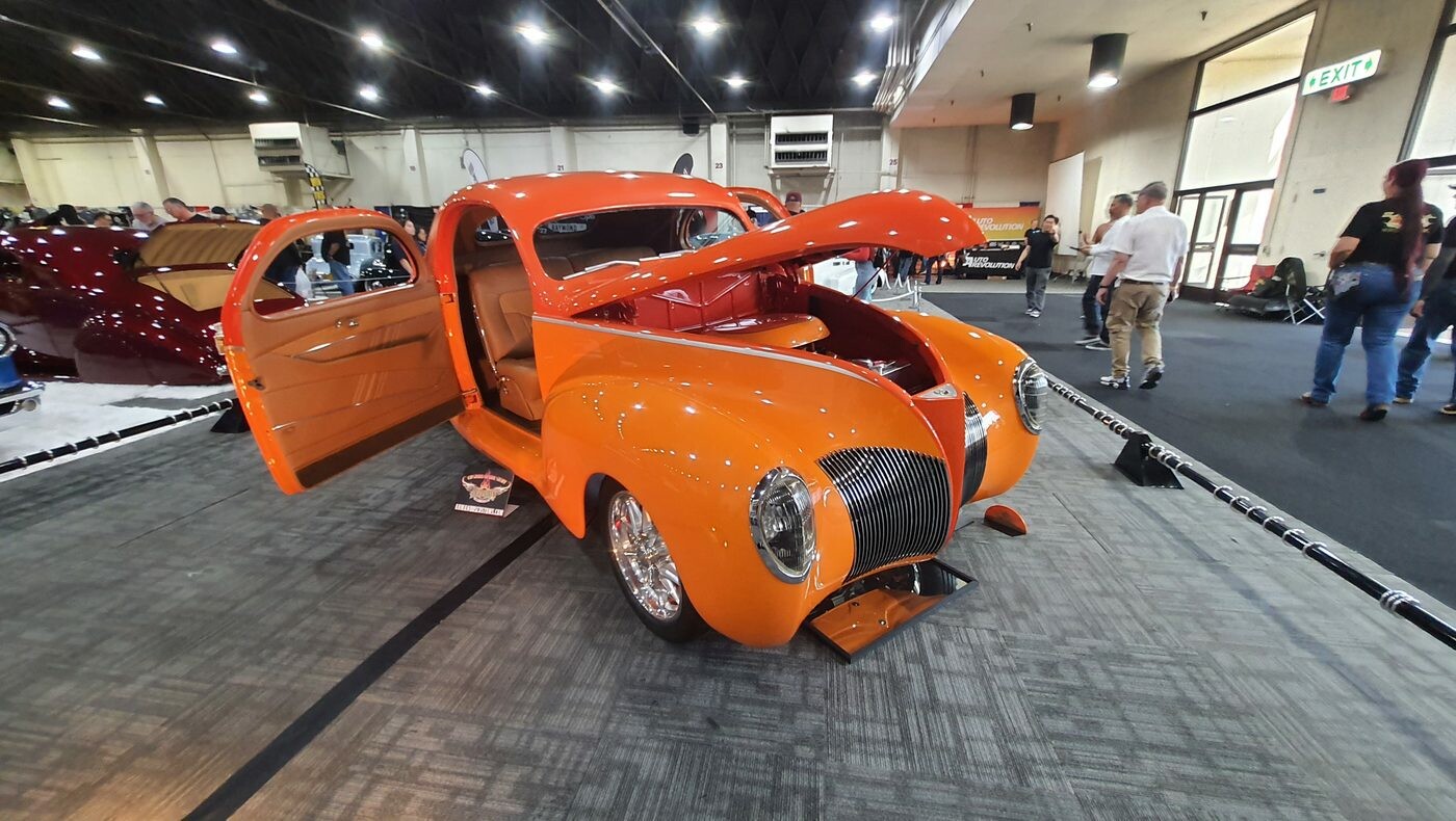 Pomarańczowy Ford Deluxe coupe z 1940 roku z otwartymi drzwiami i maską – street rod w candy na hali zlotu
