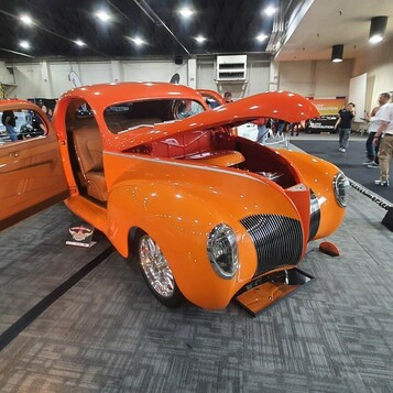 Pomarańczowy Ford Deluxe coupe z 1940 roku z otwartymi drzwiami i maską – street rod w candy na hali zlotu