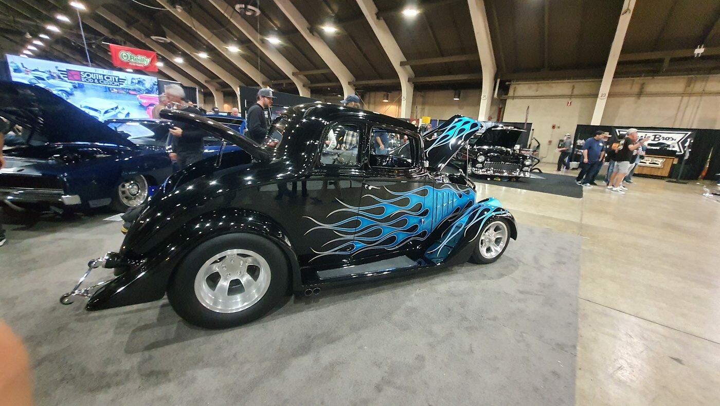 Czarny Ford 3-window coupe z 1934 roku w niebieskich płomieniach – chopped hot rod na polerowanych felgach w hali