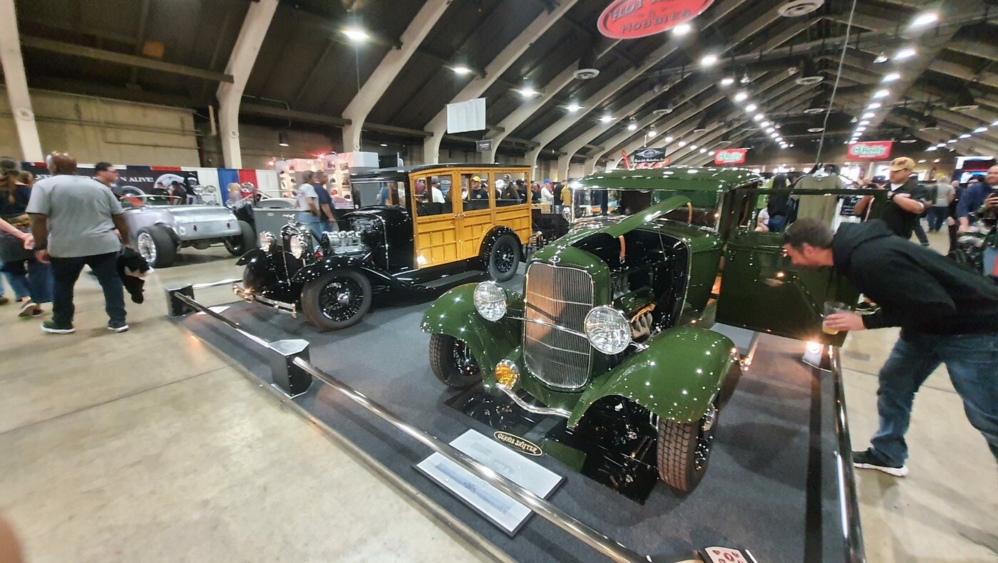 Zielony Ford 5-window coupe z 1932 roku w pełnych błotnikach obok woodie – Deuce w kolorze lasu na podeście w hali